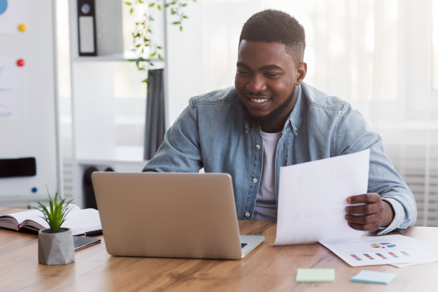 A student sits at a table in his home holding a paper and looking at a laptop