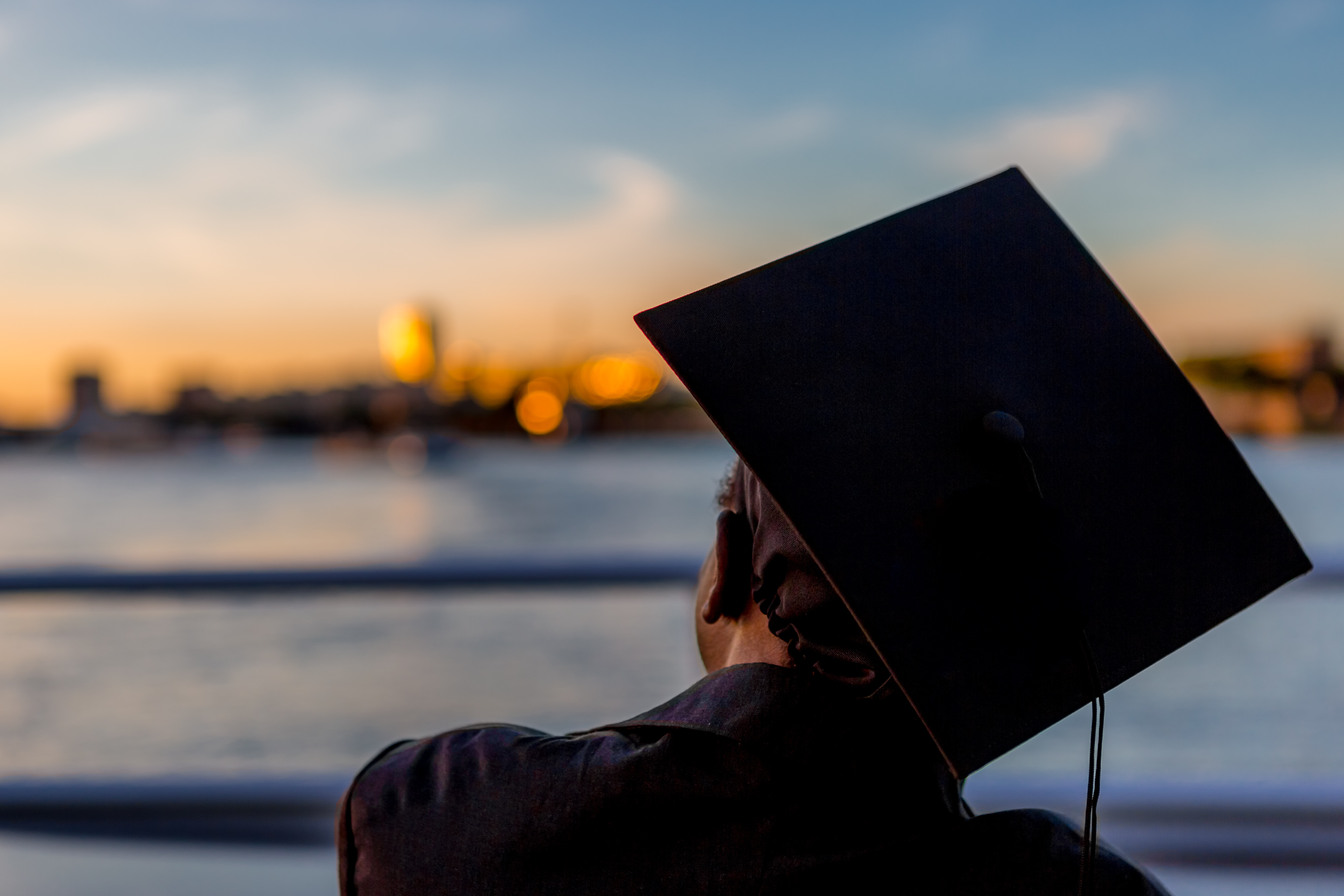 A recent graduate still wearing their cap and gownstands at a peer looking out over the water with a city in the distance..